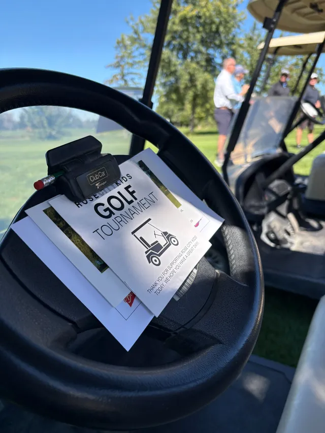 Close-up of the Rose City Kids Golf Tournament booklet clipped to a golf-cart steering wheel, with golfers standing in the background.