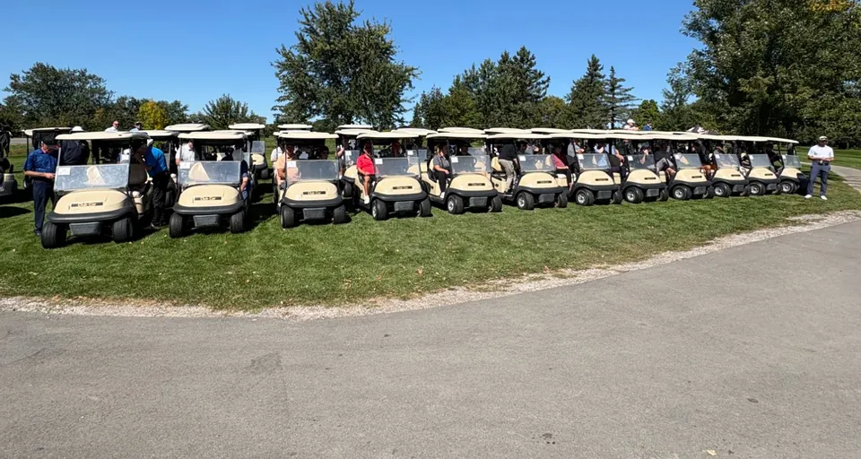 A long line of golf carts filled with tournament participants, assembled on the grass at Riverview Golf Club before the start of play.
